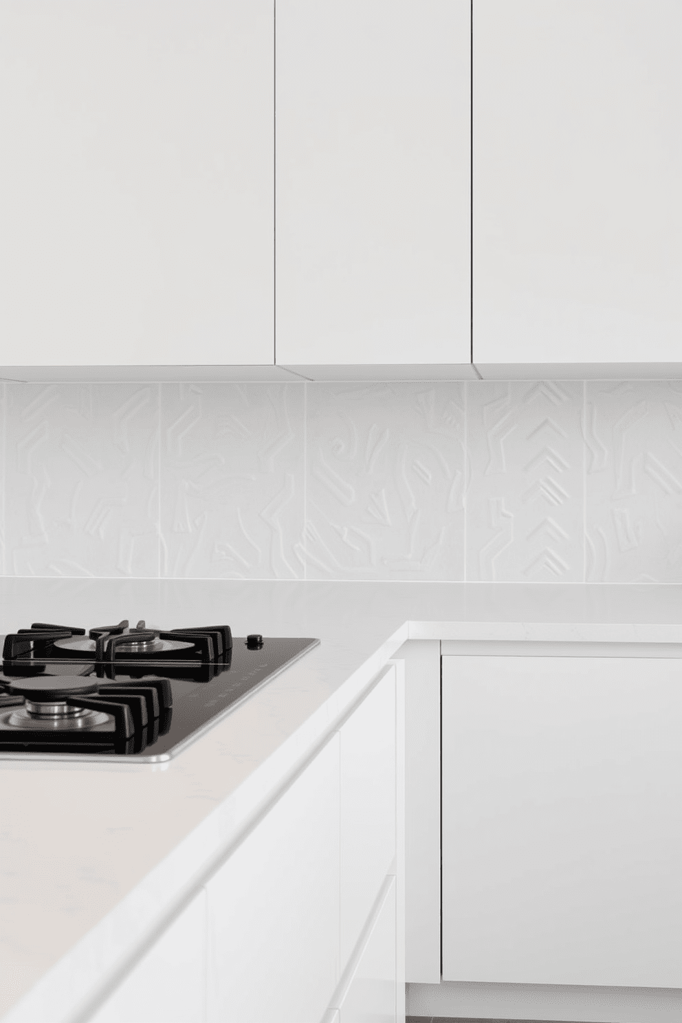 A minimalist kitchen design featuring an integrated stovetop that seamlessly blends into the countertop. The cabinetry is sleek and handleless, finished in a soft matte white, while the backsplash is a simple, textured tile in a light gray tone.